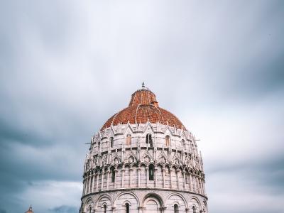 A low-angle photograph of the Pisa Baptistery of St. John, a large circular Romanesque building with white and grey striped marble walls, a terracotta-tiled dome, and a prominent central entrance, situated on a green lawn under a cloudy sky.