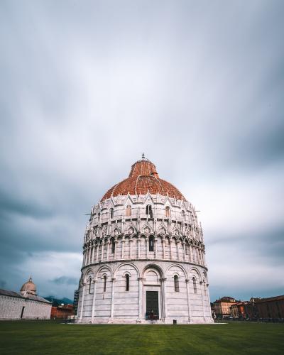 A low-angle photograph of the Pisa Baptistery of St. John, a large circular Romanesque building with white and grey striped marble walls, a terracotta-tiled dome, and a prominent central entrance, situated on a green lawn under a cloudy sky.