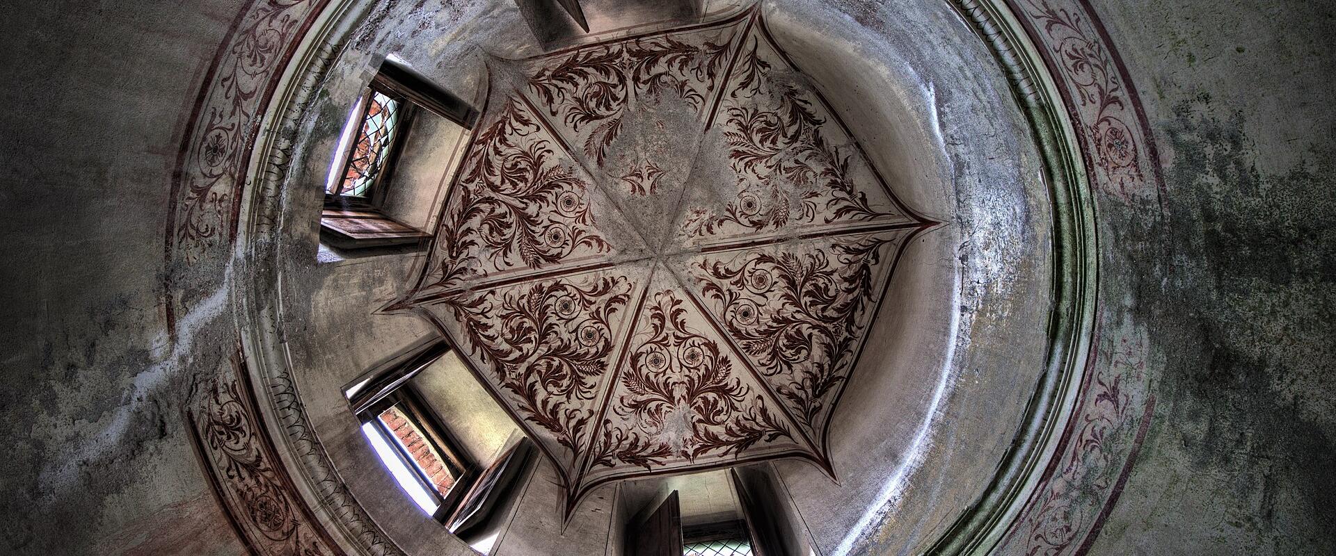 A low-angle view looking up into a circular, domed ceiling with ornate, faded floral frescoes and a central star-like painted design, surrounded by arched windows letting in daylight.