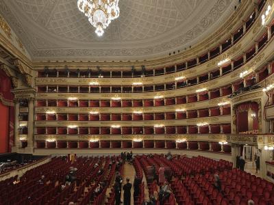A wide-angle interior view of a grand, horseshoe-shaped opera house featuring multiple tiers of ornate gold-trimmed balconies with red velvet curtains and seating, a large red stage curtain, and a central crystal chandelier.