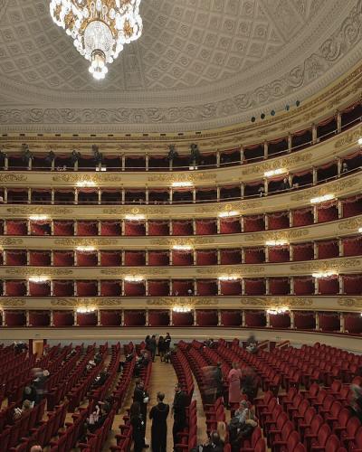 A wide-angle interior view of a grand, horseshoe-shaped opera house featuring multiple tiers of ornate gold-trimmed balconies with red velvet curtains and seating, a large red stage curtain, and a central crystal chandelier.