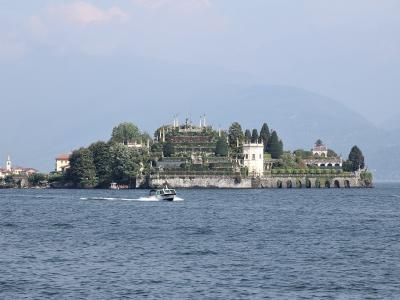 A wide shot of an island featuring a prominent Baroque palace and elaborate terraced gardens rising from the water, with a small boat passing in the foreground and a town shoreline visible to the left.