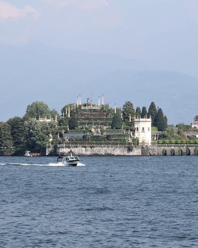A wide shot of an island featuring a prominent Baroque palace and elaborate terraced gardens rising from the water, with a small boat passing in the foreground and a town shoreline visible to the left.