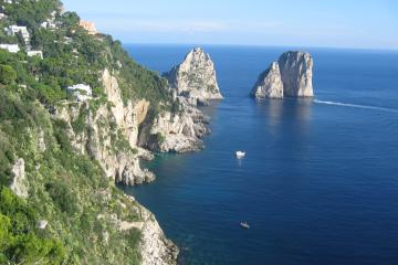 A coastal view of steep, green-covered cliffs descending to the sea, with two prominent rock formations (Faraglioni) visible offshore and a few boats on the water.