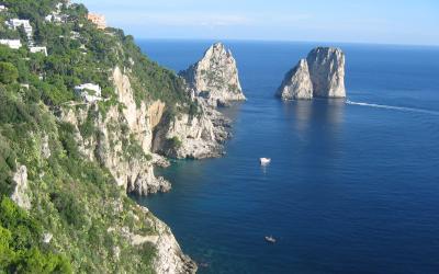A coastal view of steep, green-covered cliffs descending to the sea, with two prominent rock formations (Faraglioni) visible offshore and a few boats on the water.
