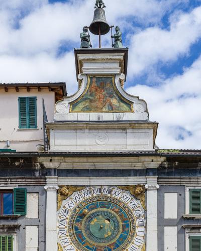 The image shows the ornate clock face and upper tower structure of the Torre dell'Orologio in Brescia, Italy, featuring a complex astronomical dial with zodiac signs, Roman numerals, golden winged figures, and two bronze statues striking a bell at