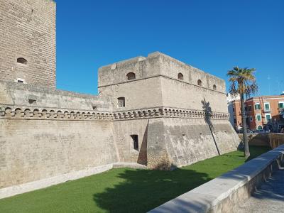 The image shows the massive stone ramparts and bastions of a medieval fortress, specifically the Castello Svevo in Bari, Italy, characterized by its thick defensive walls, crenellations, and a palm tree in the foreground.