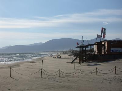 A wide sandy beach with gentle waves, a wooden lifeguard station and beach bar labeled "Base 41" on the right, roped-off sand in the foreground, and distant mountains under a partly cloudy sky.