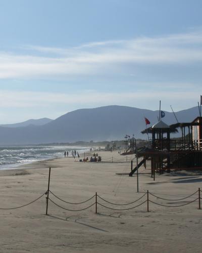 A wide sandy beach with gentle waves, a wooden lifeguard station and beach bar labeled "Base 41" on the right, roped-off sand in the foreground, and distant mountains under a partly cloudy sky.