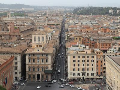 A high-angle panoramic view of a dense urban streetscape in Rome, Italy, featuring a long, straight thoroughfare (Via del Corso) lined with historic buildings and traffic, with the dome of the Pantheon visible in the distance.
