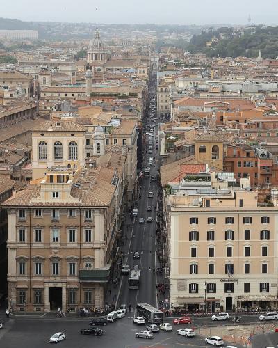 A high-angle panoramic view of a dense urban streetscape in Rome, Italy, featuring a long, straight thoroughfare (Via del Corso) lined with historic buildings and traffic, with the dome of the Pantheon visible in the distance.