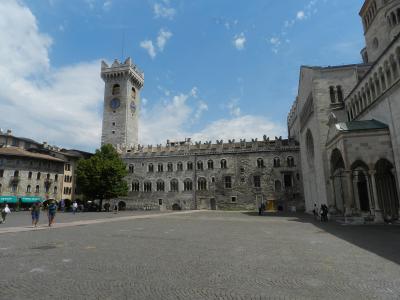 A wide-angle view of Piazza Duomo in Trento, Italy, featuring the stone tower of Palazzo dei Principi Vescovi on the left, the facade and bell tower of the Duomo di Trento (Cathedral of San Vigilio) on the right, and a paved square with pedestrian