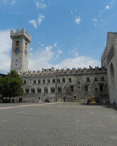 A wide-angle view of Piazza Duomo in Trento, Italy, featuring the stone tower of Palazzo dei Principi Vescovi on the left, the facade and bell tower of the Duomo di Trento (Cathedral of San Vigilio) on the right, and a paved square with pedestrian