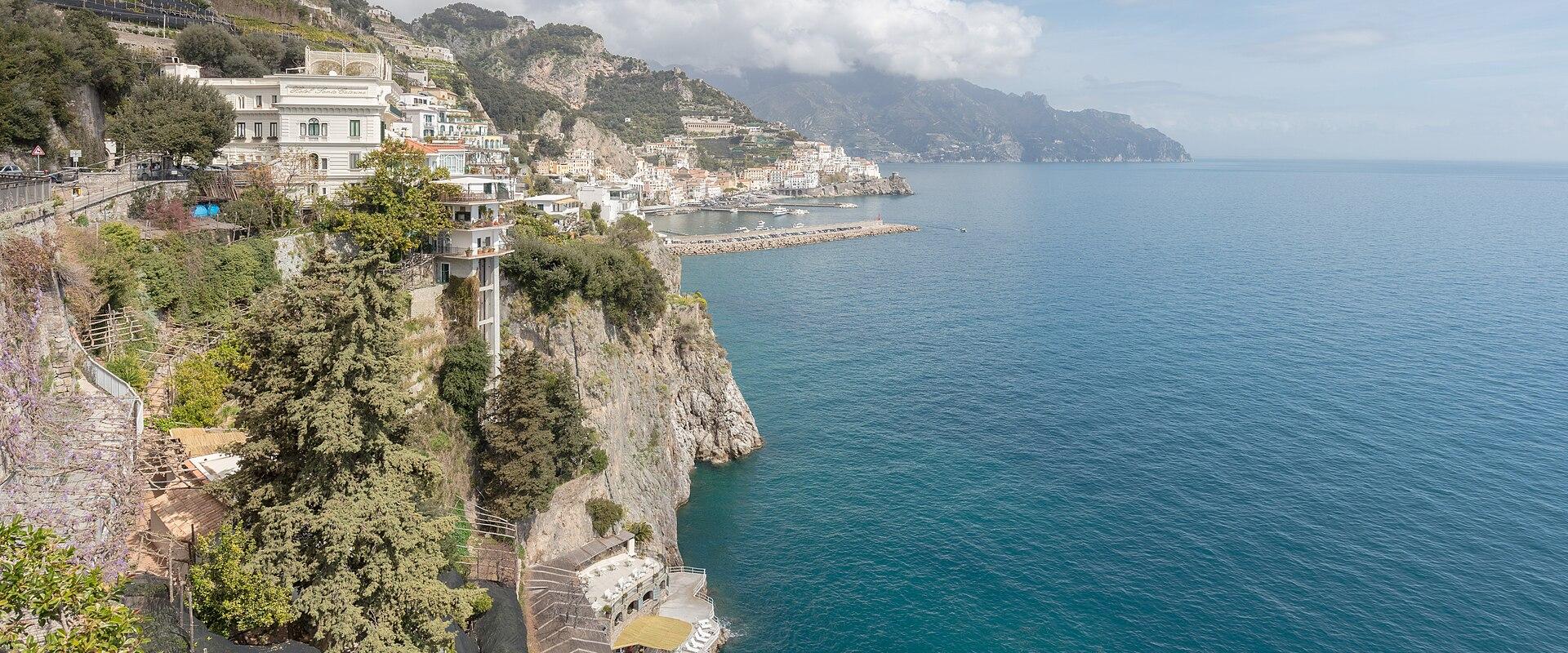 A high-angle view of a coastal town built into steep cliffs overlooking the sea, with white buildings, terraced gardens, and a small harbor visible in the distance under a partly cloudy sky.