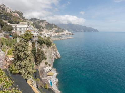 A high-angle view of a coastal town built into steep cliffs overlooking the sea, with white buildings, terraced gardens, and a small harbor visible in the distance under a partly cloudy sky.