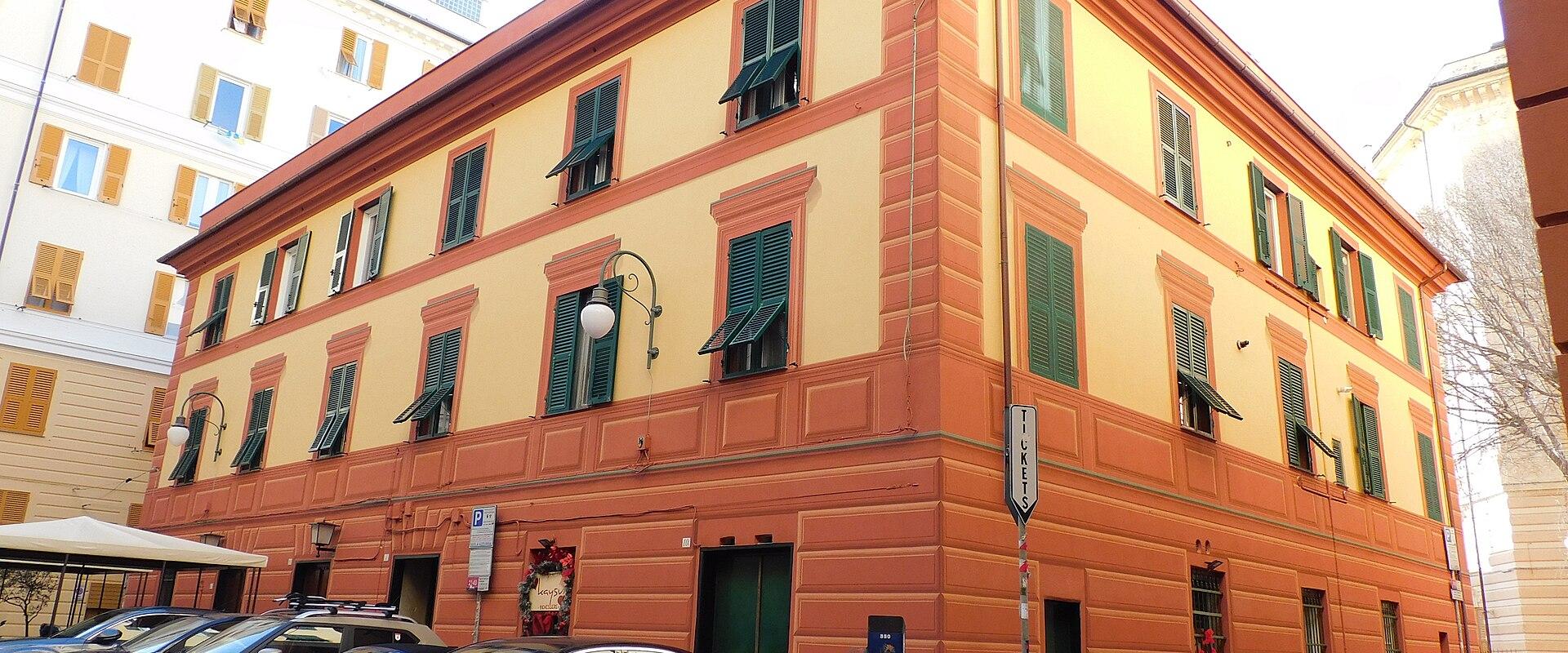 A street-level view of a large, multi-story building with ochre and terracotta facade, green shutters, parked cars, and Italian-style traffic signs in an urban European setting.