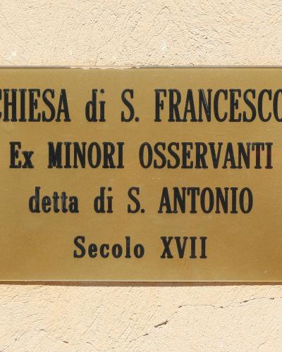 A brass plaque mounted on a textured beige wall, reading "CHIESA di S. FRANCESCO / Ex MINORI OSSERVANTI / detta di S. ANTONIO / Secolo XVII".