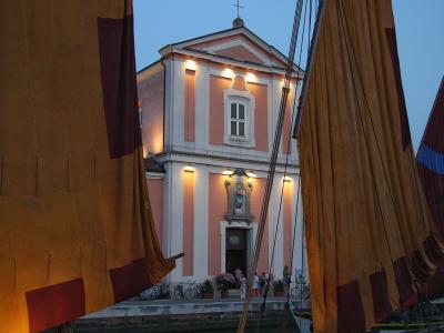 A pink and white church facade illuminated at dusk, framed by large brown sails with red trim, with people gathered at the entrance and bicycles parked nearby.
