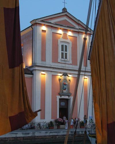 A pink and white church facade illuminated at dusk, framed by large brown sails with red trim, with people gathered at the entrance and bicycles parked nearby.