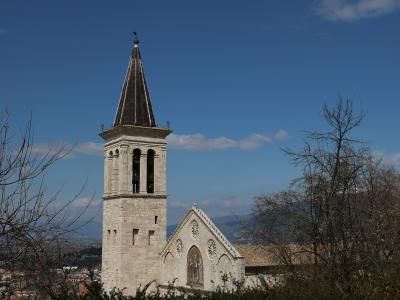 A tall, square Romanesque bell tower with a dark pyramidal spire and a church facade featuring three rose windows and a central mosaic, set against a blue sky with distant hills.