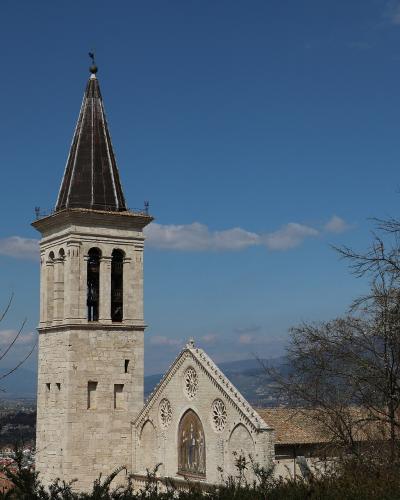 A tall, square Romanesque bell tower with a dark pyramidal spire and a church facade featuring three rose windows and a central mosaic, set against a blue sky with distant hills.