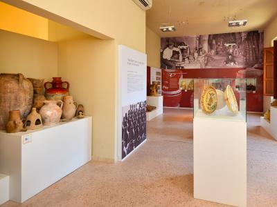 The image shows the interior of a museum gallery featuring ceramic artifacts, including large terracotta amphorae on the left and painted ceramic plates in a glass display case on the right, with informational panels and a historical photograph on