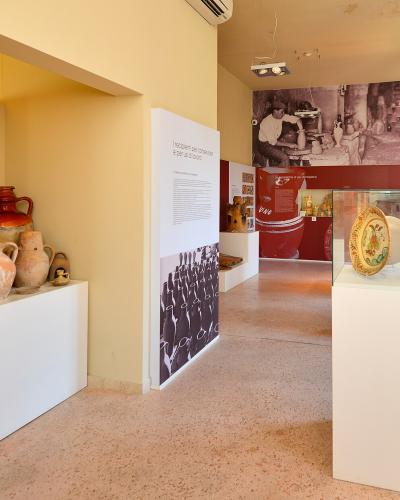The image shows the interior of a museum gallery featuring ceramic artifacts, including large terracotta amphorae on the left and painted ceramic plates in a glass display case on the right, with informational panels and a historical photograph on