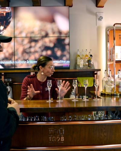 A man and a woman are at a wooden tasting bar with copper accents, where the woman is gesturing during a tasting session; the bar front and back shelves display "POLI 1898" branding, and a screen behind them shows Italian text.
