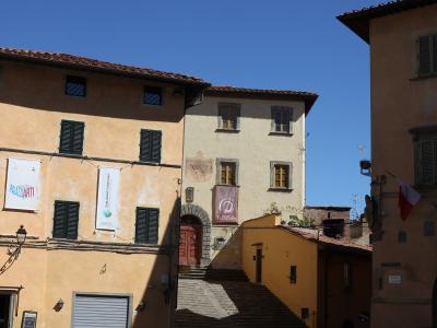 The image shows a sunlit street scene in an Italian town featuring ochre and yellow buildings with green shutters, banners for "PALAZZI D'ARTI" and "ESPLORANDO FUCECCHIO," and a small arched doorway leading up stone steps.