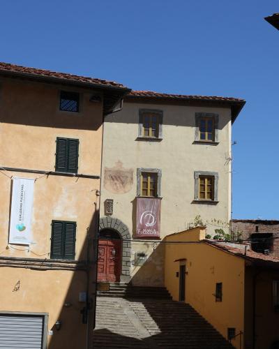 The image shows a sunlit street scene in an Italian town featuring ochre and yellow buildings with green shutters, banners for "PALAZZI D'ARTI" and "ESPLORANDO FUCECCHIO," and a small arched doorway leading up stone steps.