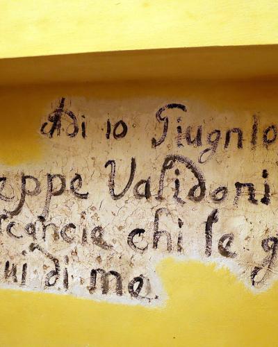 The image shows a close-up of an old, handwritten inscription on a yellow-painted wall, with text reading "Di 10 Giugno 1793 / Giuseppe Validoni Feci / queste scancie chi le guasta / ricordatevi di me." (On June 10, 1793 / Giuseppe Validoni made /