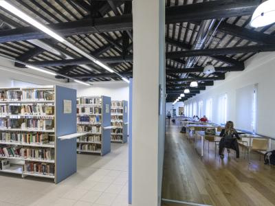 A split-view interior of a library featuring bookshelves on the left and a long reading room with wooden floors and dark ceiling beams on the right.