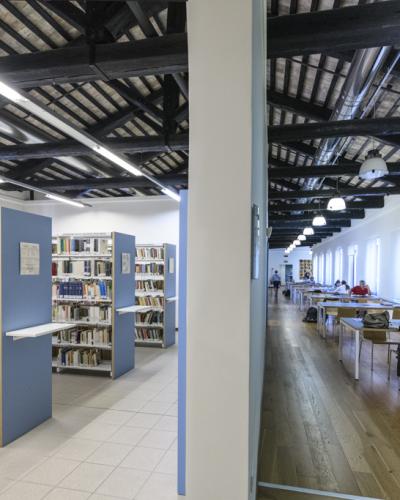 A split-view interior of a library featuring bookshelves on the left and a long reading room with wooden floors and dark ceiling beams on the right.