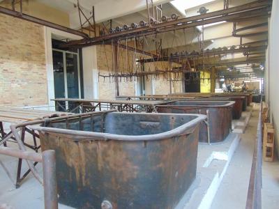 The image shows the interior of an industrial facility with large, rusted metal vats or tanks arranged in rows, overhead rail systems with hooks, and brick walls, consistent with a former slaughterhouse or abattoir.