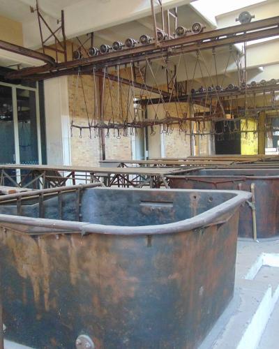 The image shows the interior of an industrial facility with large, rusted metal vats or tanks arranged in rows, overhead rail systems with hooks, and brick walls, consistent with a former slaughterhouse or abattoir.