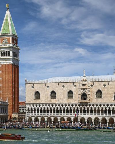 The image shows the Doge’s Palace (Palazzo Ducale) and St. Mark’s Campanile in Venice, Italy, viewed from the water with boats and gondolas in the foreground under a partly cloudy sky.