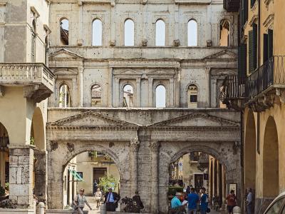 A low-angle view of the ancient Roman Porta Borsari in Verona, Italy, showing its two lower arches with pedestrians and cyclists passing through, flanked by historic buildings, with the upper levels of the gate featuring rows of arched windows aga