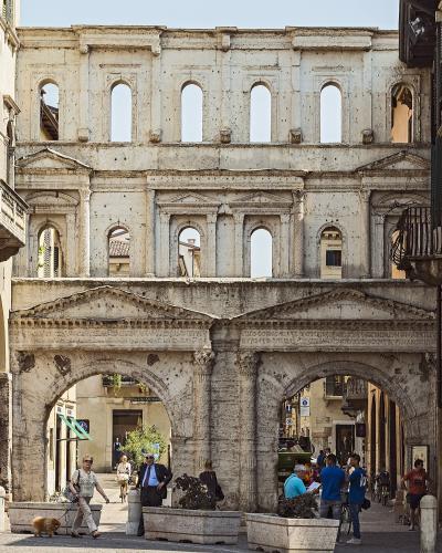 A low-angle view of the ancient Roman Porta Borsari in Verona, Italy, showing its two lower arches with pedestrians and cyclists passing through, flanked by historic buildings, with the upper levels of the gate featuring rows of arched windows aga