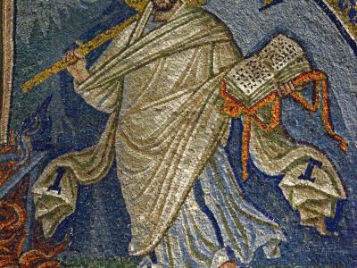 A close-up photograph of a Byzantine-style mosaic depicting Saint Lawrence, identifiable by the gridiron (grill) he holds and the flames at his feet, set against a blue background with gold tesserae.