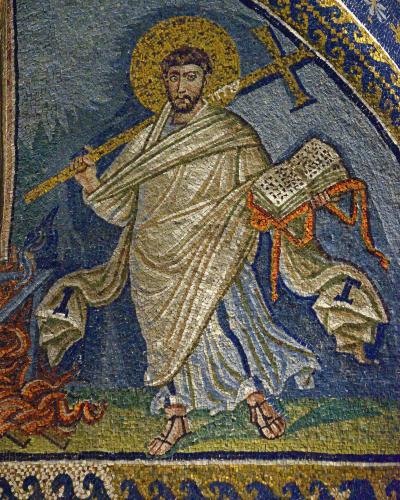 A close-up photograph of a Byzantine-style mosaic depicting Saint Lawrence, identifiable by the gridiron (grill) he holds and the flames at his feet, set against a blue background with gold tesserae.