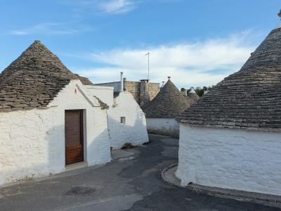 The image shows a cluster of traditional white-washed stone houses with conical roofs made of grey slate, characteristic of trulli architecture, set along a paved street under a blue sky with scattered clouds.