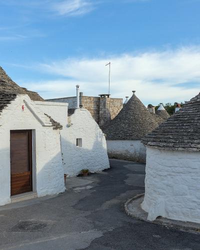 The image shows a cluster of traditional white-washed stone houses with conical roofs made of grey slate, characteristic of trulli architecture, set along a paved street under a blue sky with scattered clouds.