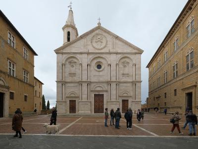 The image shows the facade of a Renaissance-style cathedral with a central bell tower, situated in a paved town square flanked by two large historic buildings, with several people and a dog visible in the foreground.