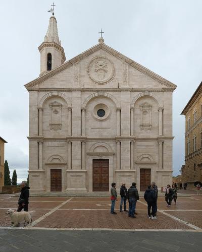 The image shows the facade of a Renaissance-style cathedral with a central bell tower, situated in a paved town square flanked by two large historic buildings, with several people and a dog visible in the foreground.