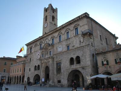 A large, historic stone palace with a prominent clock tower, arched ground-floor loggias, and flags flying on the left side, situated in a paved public square with people and outdoor café umbrellas.