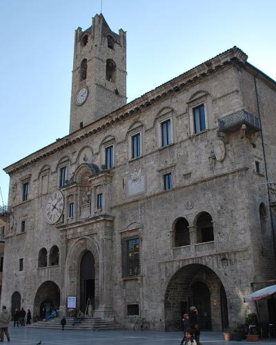 A large, historic stone palace with a prominent clock tower, arched ground-floor loggias, and flags flying on the left side, situated in a paved public square with people and outdoor café umbrellas.