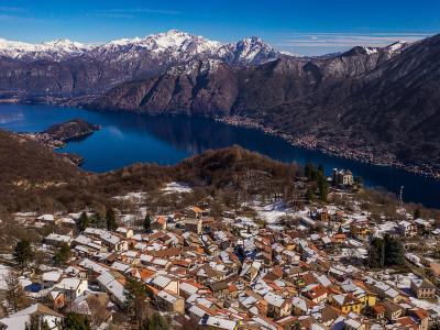 A high-angle panoramic view of a snow-dusted hillside village overlooking Lake Como, with the town of Brunate visible in the foreground and the lake and distant Alps in the background.
