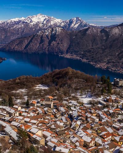 A high-angle panoramic view of a snow-dusted hillside village overlooking Lake Como, with the town of Brunate visible in the foreground and the lake and distant Alps in the background.