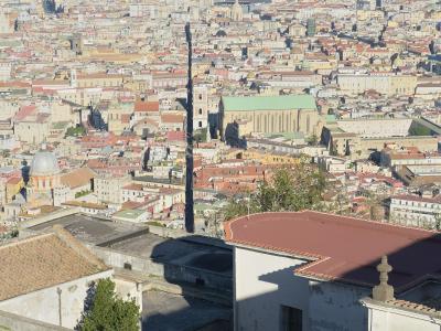A high-angle panoramic view of the city of Naples, Italy, looking south from an elevated position (likely Castel Sant'Elmo), showing the dense urban fabric, the large green-roofed Duomo (Naples Cathedral), and the distant mountains.