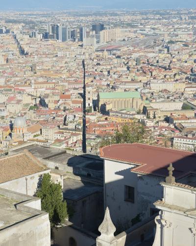 A high-angle panoramic view of the city of Naples, Italy, looking south from an elevated position (likely Castel Sant'Elmo), showing the dense urban fabric, the large green-roofed Duomo (Naples Cathedral), and the distant mountains.
